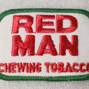 May include: Embroidered patch with the words "RED MAN" in red capital letters above "CHEWING TOBACCO". The patch has a white background with a green border.