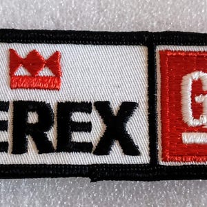 May include: Embroidered patch with the word "TEREX" in black block letters, a red crown, and a red square with "GM" in white. The patch has a white background and a black border.