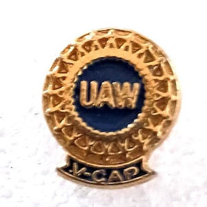 May include: A gold-toned UAW pin with a blue center. The pin features the letters "UAW" in gold against a blue background, surrounded by a decorative gold border. The bottom of the pin reads "V-CAP" in gold.
