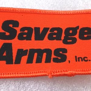 May include: Rectangular orange patch with the words "Savage Arms, Inc." in bold black letters. The patch has a stitched orange border.