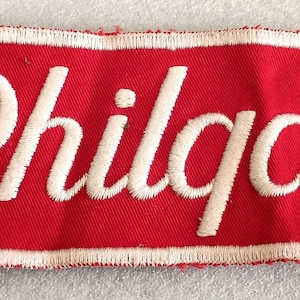 May include: A rectangular red fabric patch with the word "Philgas" embroidered in white cursive script. The patch has a white border and is likely an emblem or logo.