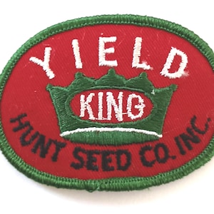 May include: An oval-shaped patch with a red background and a green border. The patch features the words "YIELD KING" in white, with "KING" inside a green crown. Below, it reads "HUNT SEED CO. INC." in black.