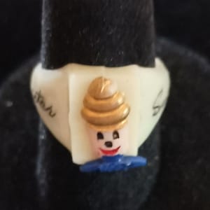 May include: A cream-colored ring with a small, smiling clown figure. The clown has a gold-colored hat, a red mouth, and a blue collar. The ring is on a black surface.