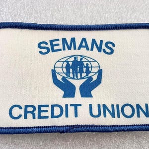May include: A rectangular patch with a white background and a blue border. The patch features the words "SEMANS CREDIT UNION" in blue, along with a graphic of a globe held by two hands, with silhouettes of people inside.