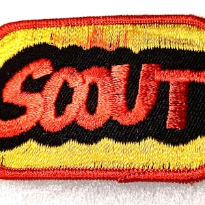May include: Embroidered patch with the word "SCOUT" in red, outlined in black, on a black background. The top and bottom of the patch have a yellow fringe design. The patch is rectangular with a red border.