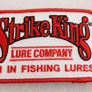 May include: Embroidered patch with a white background and a red border. The patch features the words "Strike King" in red, stylized lettering, above "LURE COMPANY" and "#1 IN FISHING LURES" in red.