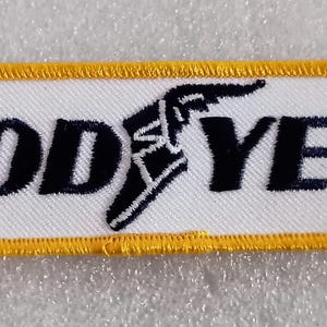 May include: A rectangular patch with the word "GOOD YEAR" in bold black letters. A black and white winged foot logo is between the words. The patch has a white background with a yellow border.