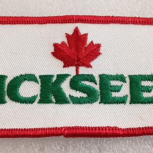 May include: Embroidered patch with a white background, red border, and the word "PICKSEED" in green. A red maple leaf is centered above the text. The patch is rectangular.