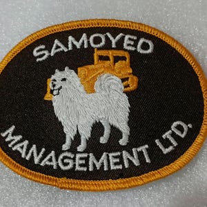 May include: Embroidered patch with a white Samoyed dog and a yellow bulldozer on a black background. The patch is oval-shaped with the words "SAMOYED MANAGEMENT LTD." in white lettering and a gold border.