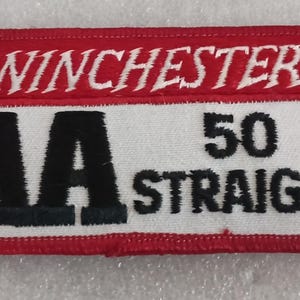 May include: A rectangular patch with a red border and background. The top section has the word "WINCHESTER" in white script. Below, a white section displays "AA 50 STRAIGHT" in black block letters.
