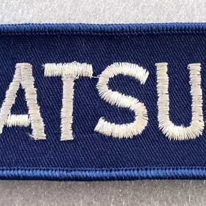 May include: A rectangular patch with the word "DATSUN" embroidered in white thread on a navy blue background. The patch has a navy blue border with white stitching.