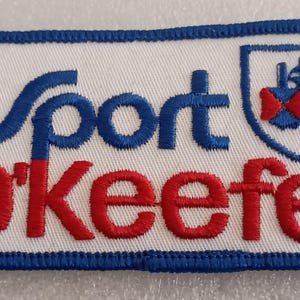 May include: Embroidered patch with the words "Sport O'Keefe" in blue and red lettering. The patch has a white background with a blue border and a shield emblem in the upper right corner.