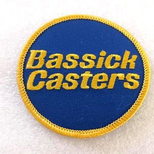 May include: Round blue patch with yellow embroidered text that reads "Bassick Casters". The patch has a yellow embroidered border. The patch is likely for a jacket or bag.