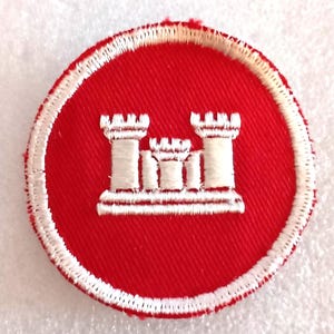 May include: A circular red patch with a white embroidered border. The patch features a white emblem of three castle turrets. This patch is likely a military or organizational insignia.