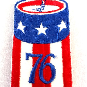 May include: Embroidered patch in red, white, and blue, resembling a firecracker. The top section is blue with white stars, the middle has red and white stripes, and the number "76" is in blue. A small red string extends from the top.