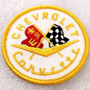 May include: A round, white patch with the word "CHEVROLET" arched across the top in gold lettering. The word "Corvette" is arched across the bottom. Two crossed flags, one red and one black and white checkered, are in the center.