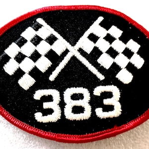 May include: An oval-shaped embroidered patch with a black background and a red border. The design features two crossed white checkered racing flags positioned above the white embroidered number 383, centered on the black fabric surface.