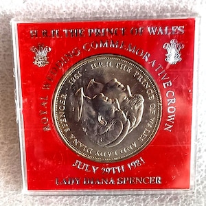 May include: A commemorative coin from the Royal Wedding of Prince Charles and Lady Diana Spencer, dated July 29th, 1981. The coin is silver-colored and encased in a red protective sleeve. Text includes "H.R.H. The Prince of Wales" and "Commemorative Crown."