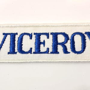 May include: A rectangular patch with the word "VICEROY" embroidered in blue capital letters on a white background. The patch has a white border with a stitched edge.