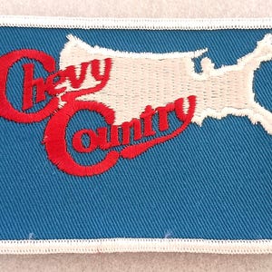 May include: A rectangular embroidered patch with a blue background and a white border. It features a white silhouette of the United States map and the red embroidered text Chevy Country, with the word Chevy above the word Country in a script font.