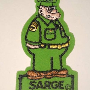 May include: Green embroidered patch featuring a cartoon character in a green uniform with a gold emblem. The patch has the word "SARGE" in black lettering at the bottom. The character has a light skin tone and a hat.