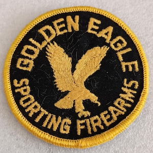 May include: Round patch with a black background and a gold eagle design. The text "GOLDEN EAGLE SPORTING FIREARMS" is in gold around the edge, with a yellow border.