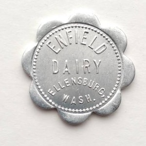 May include: A silver-colored metal token with a scalloped edge. The text "ENFIELD DAIRY ELLENSBURG WASH." is embossed in the center. The token has a circular shape with a beaded border.