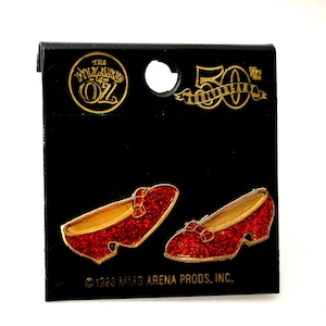 May include: A black card featuring two red glittery shoe pins with gold trim, reminiscent of Dorothy's ruby slippers from The Wizard of Oz. The card also has the text "The Wizard of Oz" and "50th Anniversary".