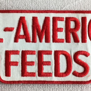 May include: Embroidered patch with the words "ALL-AMERICAN FEEDS" in red, stitched onto a white rectangular background with a red border. The patch has a unique shape with a cutout at the bottom.