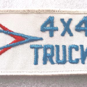 May include: Embroidered patch with the words "4X4 TRUCKS" in blue, and a red and blue arrow design on a white background. The patch has a stitched border.