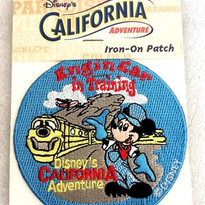 May include: A round embroidered iron-on patch featuring Mickey Mouse in blue overalls and a cap, standing next to a yellow train. The text reads "Engineer in Training" and "Disney's California Adventure" on a light blue background with clouds.