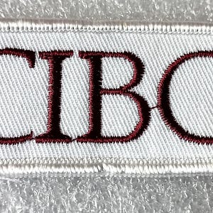 May include: A rectangular embroidered patch featuring a white background with a white stitched border. The letters CIBC are embroidered in a dark maroon thread across the center of the patch in a serif font style.