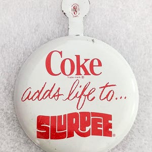 May include: Vintage white metal Coke and Slurpee advertising button. The button features the word "Coke" in red, the phrase "adds life to..." in cursive, and the word "Slurpee" in a bold red font. The button has a small handle at the top.