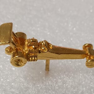 May include: A small, gold-colored pin shaped like a vintage race car. The pin features a detailed engine, a rear wing, and small wheels. The pin is designed with a single post on the back for secure attachment to clothing or accessories.