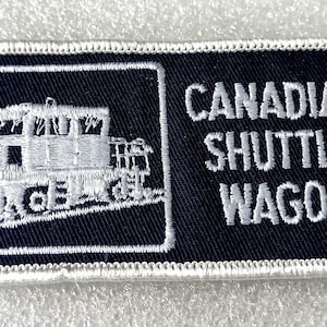May include: A rectangular embroidered patch with a black background and white border. The left side features a white stitched graphic of a train car, while the right side displays the white embroidered text CANADIAN SHUTTLE WAGON.