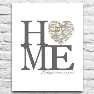 May include: A grey print with the word "HOME" in large letters. The "O" is replaced with a heart-shaped map of Raleigh, North Carolina. The text "Raleigh, North Carolina" appears below the word "HOME".