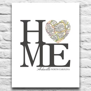 Op de afbeelding: Een zwart-witte print met het woord "HOME" in grote letters. De "O" is vervangen door een hartvormige kaart van Asheville, North Carolina. De tekst "Asheville, North Carolina" staat onder het woord "HOME".