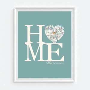 May include: A teal blue background with the word "HOME" in white letters. The "O" is replaced with a heart-shaped map of Macon, Georgia. The text "Macon GEORGIA" is written in white below the word "HOME".