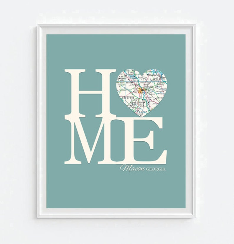 May include: A teal blue background with the word "HOME" in white letters. The "O" is replaced with a heart-shaped map of Macon, Georgia. The text "Macon GEORGIA" is written in white below the word "HOME".