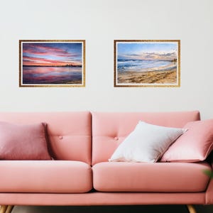 May include: Two framed landscape art prints. One depicts a sunset over water with pink, orange, and blue hues. The other shows a beach scene with waves and a blue sky. Both are in gold frames.