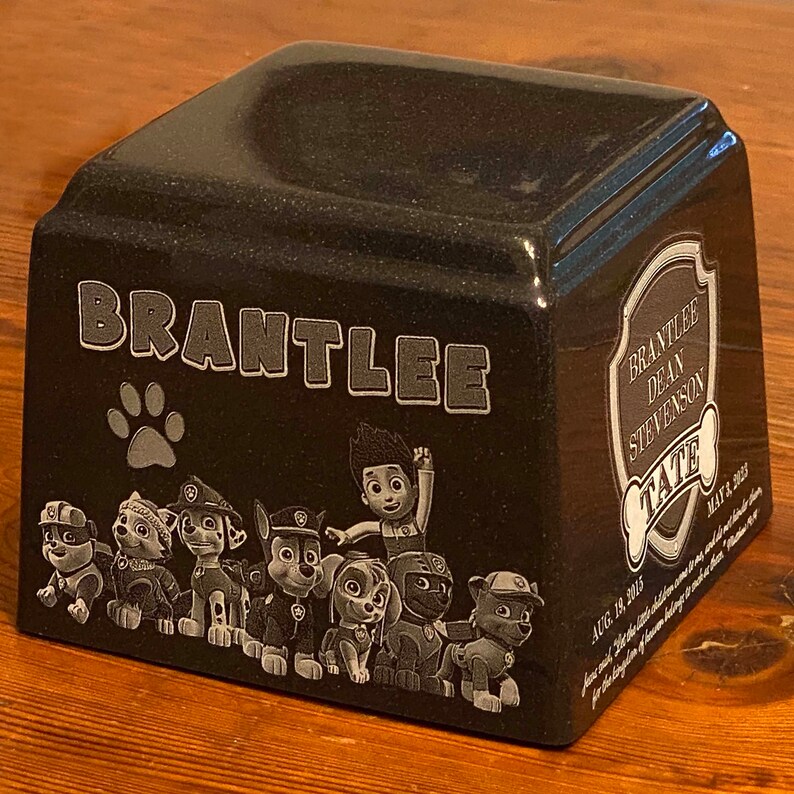 May include: A black granite pet urn with a paw print and the name 'BRANTLEE' engraved on the front. The urn also features a Paw Patrol character design and the text 'BRANTLEE DEAN STEVENSON TATE AUG. 19, 2015 MAY 18, 2023'.
