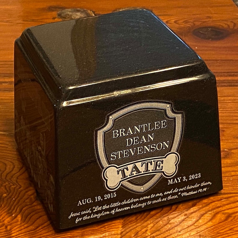 May include: A black granite pet urn with a silver shield engraved with the name 'Brantlee Dean Stevenson Tate'. The urn also has the dates 'AUG. 19, 2015' and 'MAY 3, 2023' engraved on it. The inscription below the shield reads 'Jesus said, 'Let the little children come to me, and do not hinder them, for the kingdom of heaven belongs to such as these.' Matthew 19:14'