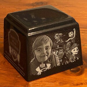 May include: A black granite urn with a photo of a young boy and cartoon characters. The urn is engraved with the text 'Brantlee Dean Stevenson', 'Tate', 'Aug 18, 2016', 'May 3, 2023', and 'In loving memory of our children who are now our guardian angels.  We love you more than words can say.  Rest in Peace.'