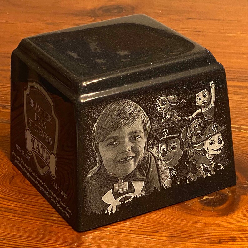 May include: A black granite urn with a photo of a young boy and cartoon characters. The urn is engraved with the text 'Brantlee Dean Stevenson', 'Tate', 'Aug 18, 2016', 'May 3, 2023', and 'In loving memory of our children who are now our guardian angels.  We love you more than words can say.  Rest in Peace.'