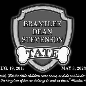 May include: A black and white memorial plaque with a shield shape and a bone on each side. The shield has the text 'BRANTLEE DEAN STEVENSON TATE' and the dates 'AUG. 19, 2015' and 'MAY 3, 2023'. The plaque also has a quote from the Bible: 'Jesus said, 'Let the little children come to me, and do not hinder them, for the kingdom of heaven belongs to such as these.' Matthew 19:14'