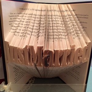 Peut inclure: Une sculpture d'art de livre avec le mot "remarquable" plié dans les pages. Le livre est ouvert et debout sur une table.