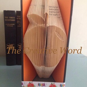 May include: A book folded into the shape of an apple with a ribbon that says "Best Teacher" around the bottom. The book is standing on a shelf with other books.