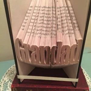May include: A book art sculpture of the word "handmade" created by folding the pages of a book. The book is standing on a red book with the title "The Bleak House" on the spine.
