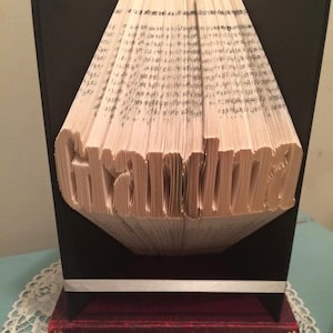 May include: A book art sculpture of the word "Grandma" folded from the pages of a book. The sculpture is displayed on a black stand and a red book with the title "The Bleak House" on the spine.