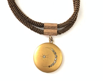 May include: A gold locket necklace with a crescent moon design and a single star in the center. The necklace is made of a brown braided cord and has a gold clasp.